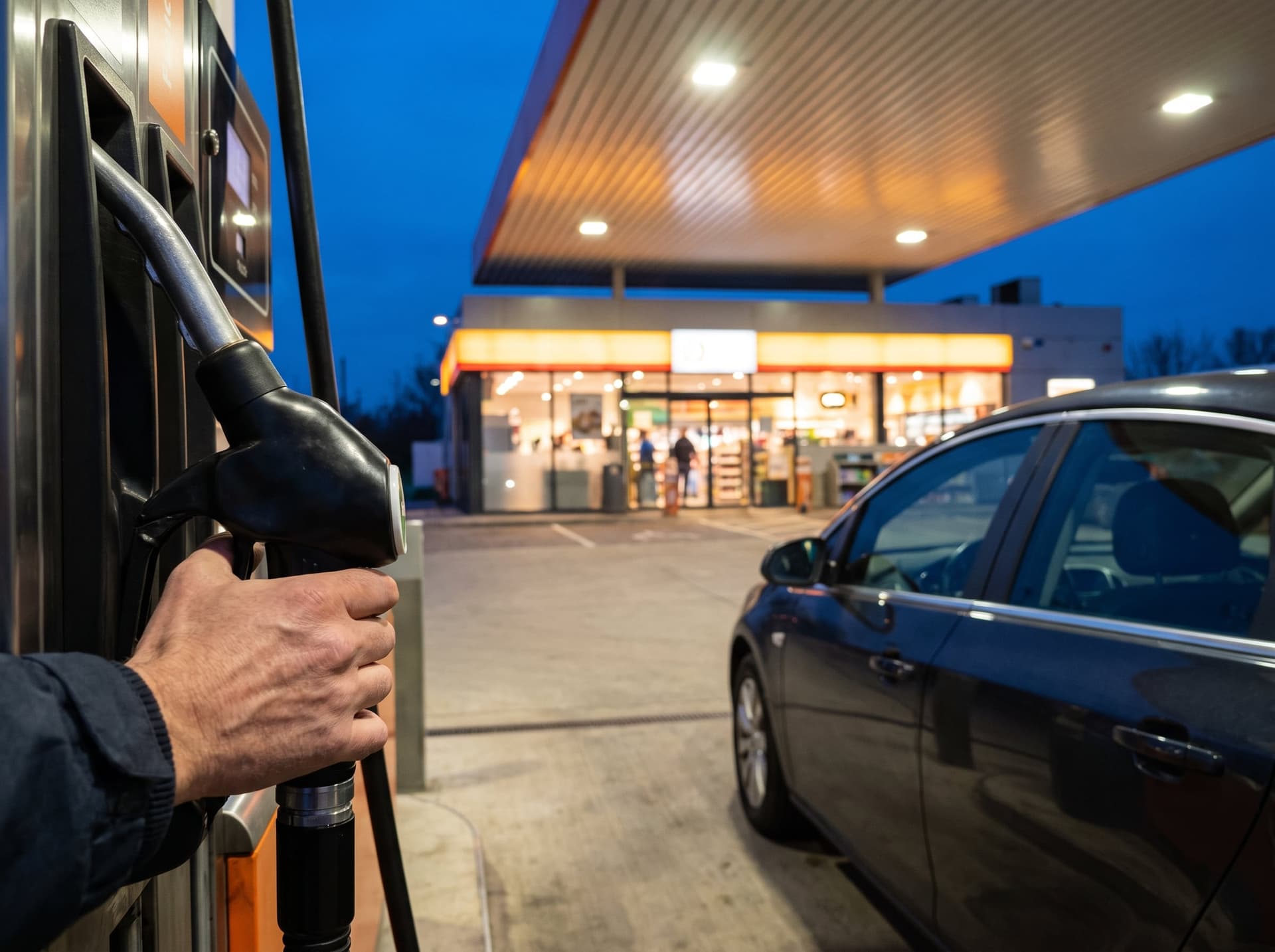 A hand lifting a fuel nozzle at a pump island under a lit canopy at dusk